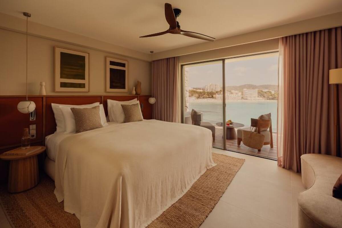 A hotel room with a large bed, a ceiling fan, a view of the sea, and a balcony with seating.