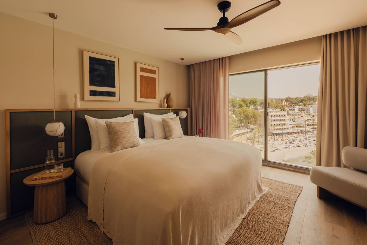 A bedroom with a large bed, a ceiling fan, a view of a beach, and a cityscape.