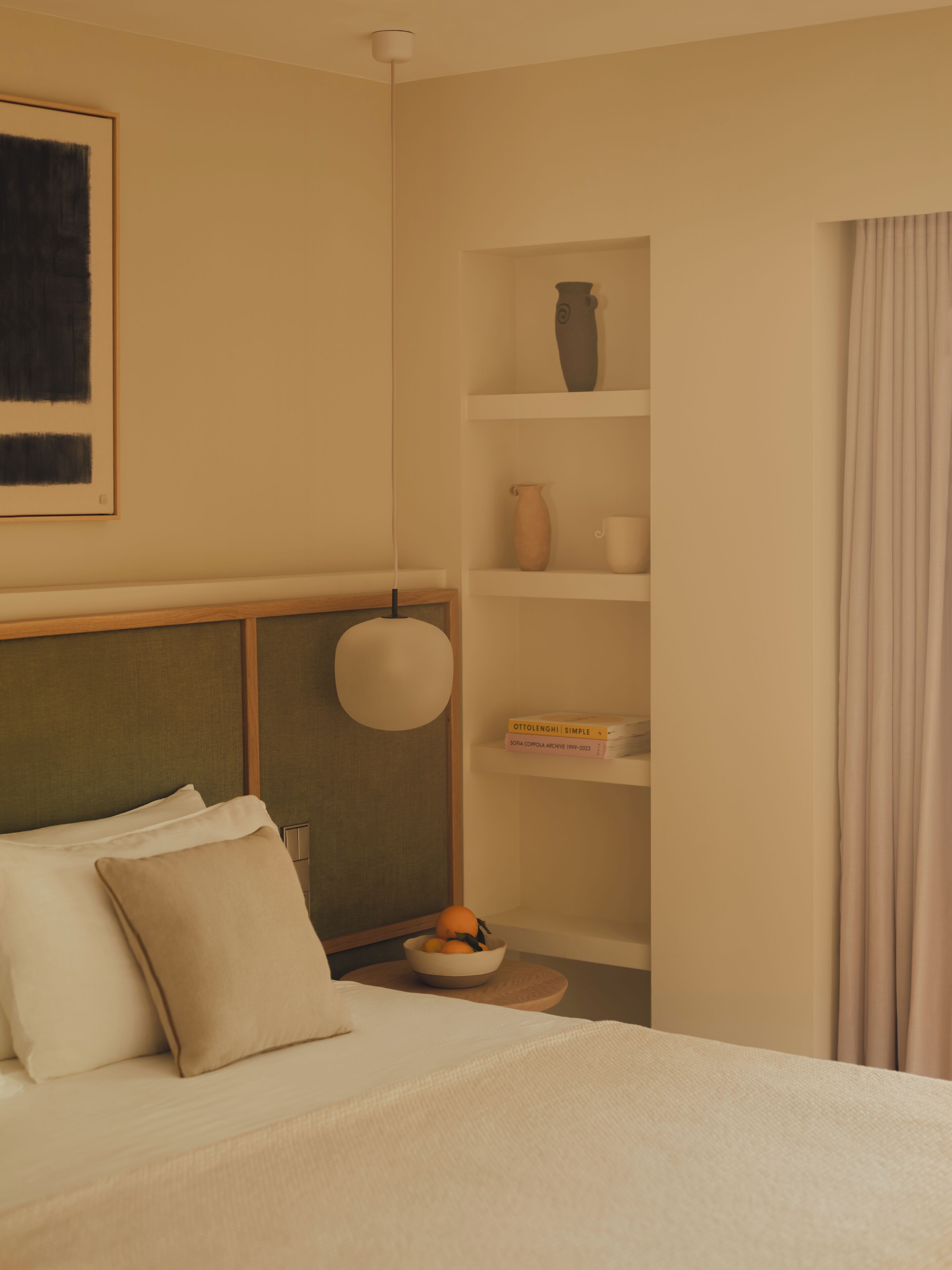 A neatly arranged hotel room with a bed, pillows, a bedside table with a bowl of fruit, a shelf with decorative items, and a hanging light fixture.