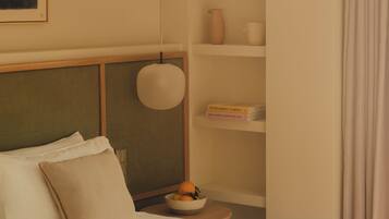 A neatly arranged hotel room with a bed, pillows, a bedside table with a bowl of fruit, a shelf with decorative items, and a hanging light fixture.