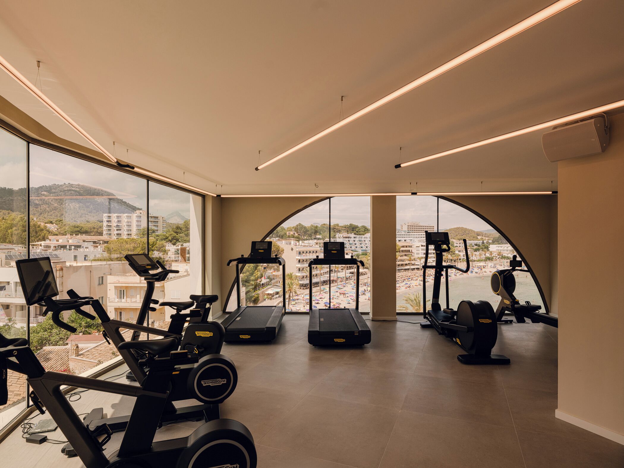 A modern gym with various exercise equipment, large windows offering views of a cityscape and beach.