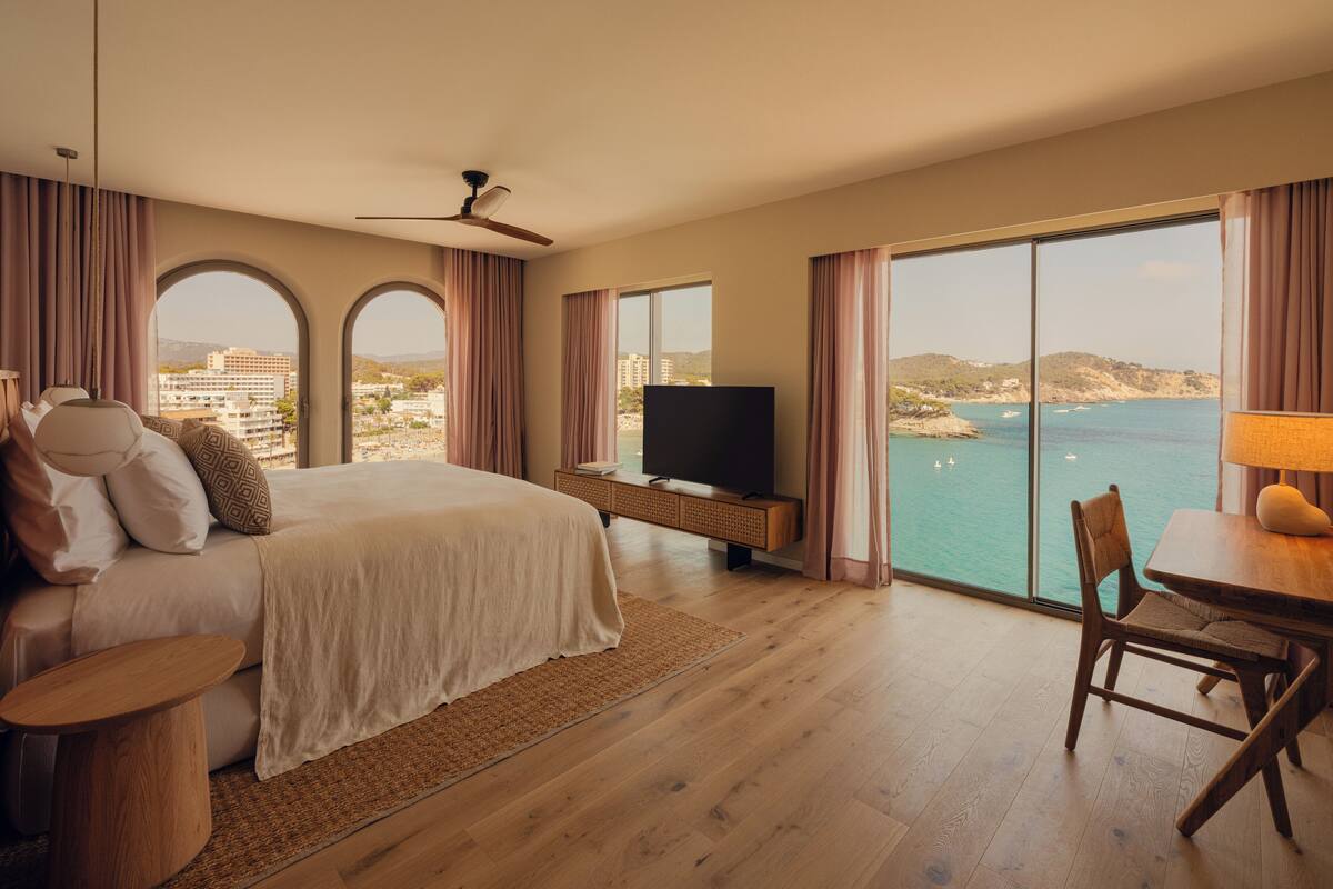 A hotel room with a large bed, a TV, a dining area, and a view of the sea.