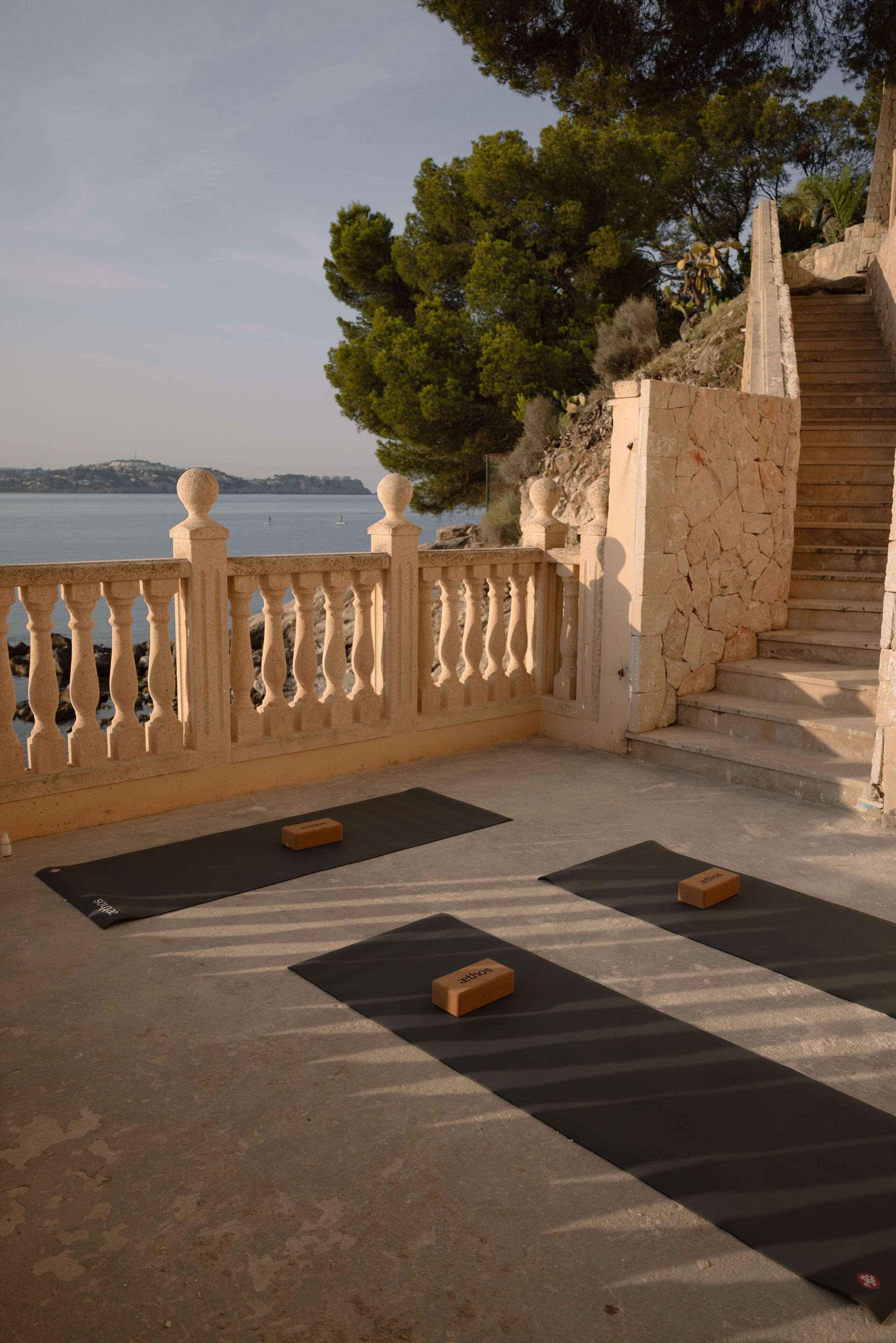 A balcony with black mats and wooden blocks, overlooking a body of water and a stone staircase.