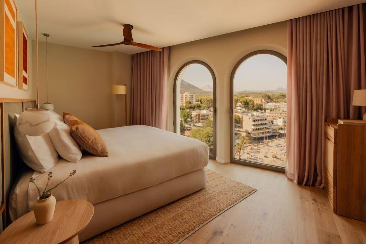 A bedroom with a large bed, a ceiling fan, a view of the city through arched windows, and a wooden nightstand.