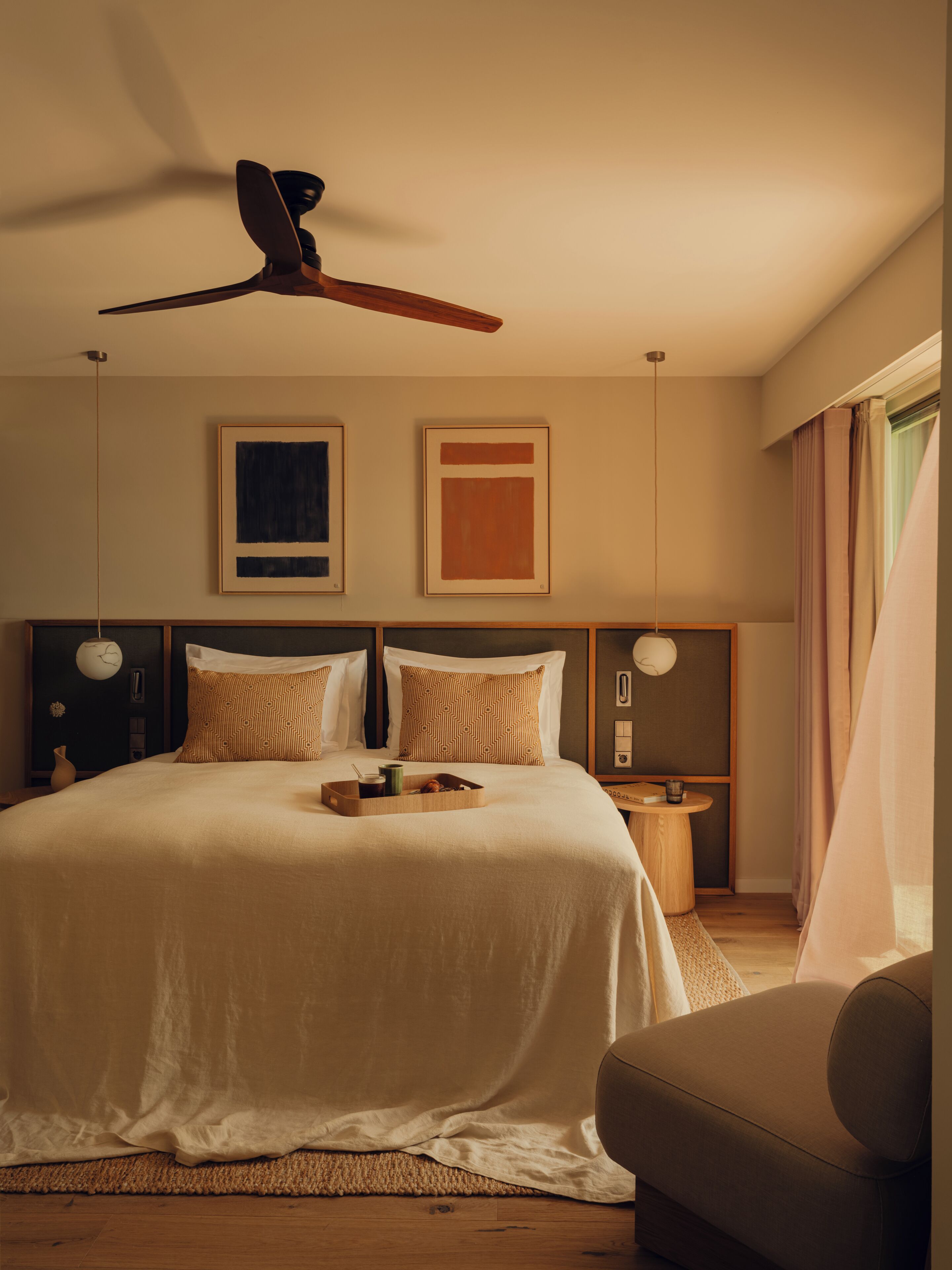 A bedroom with a bed, a ceiling fan, two framed artworks, a bedside table with a lamp, and a chair.