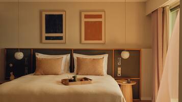 A bedroom with a bed, a ceiling fan, two framed artworks, a bedside table with a lamp, and a chair.