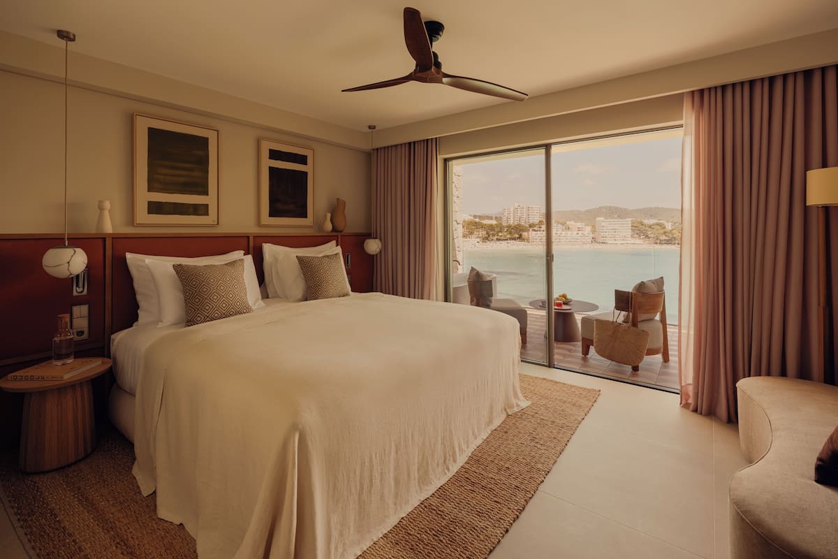 A hotel room with a large bed, a ceiling fan, a view of the beach, and a balcony with seating.