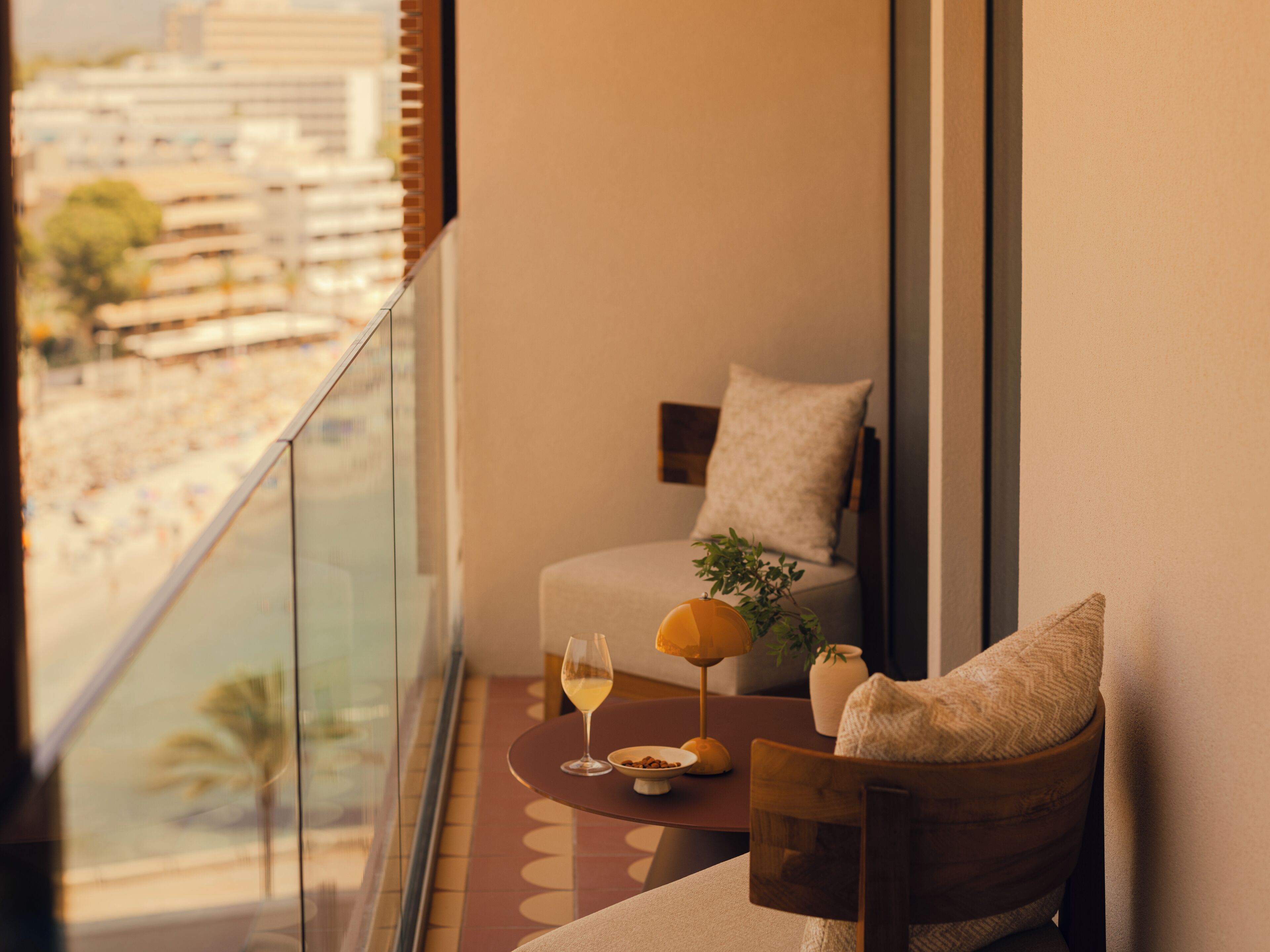 A balcony with a view of a beach and buildings, a sofa with a cushion, a small table with a glass of wine, and a plant.