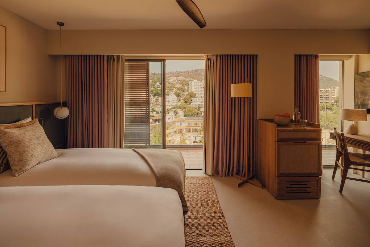 A hotel room with two beds, a desk, a chair, a ceiling fan, and a view of the city through large windows.
