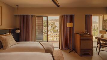 A hotel room with two beds, a desk, a chair, a ceiling fan, and a view of the city through large windows.