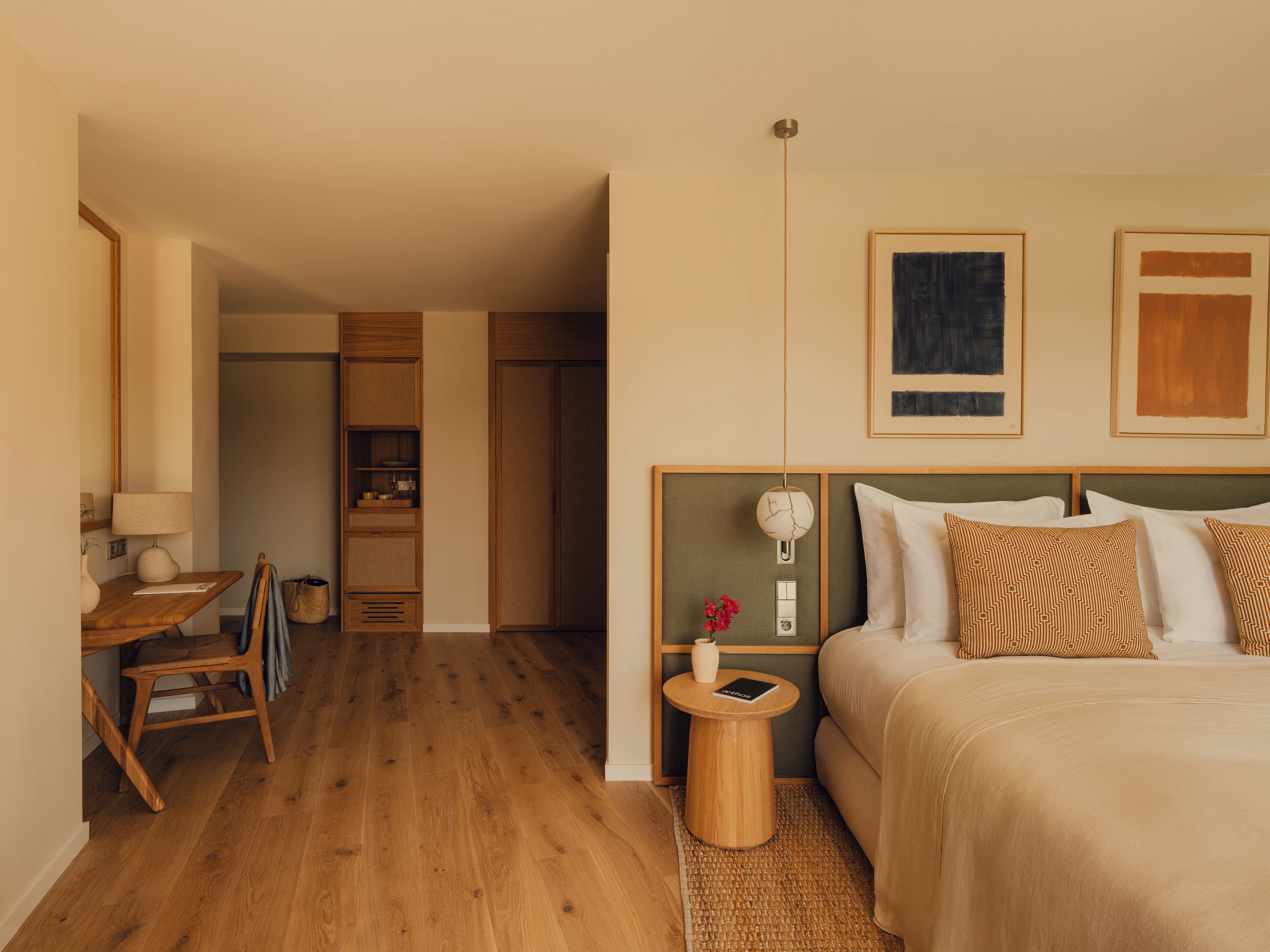 A hotel room with a wooden floor, a bed with patterned pillows, a bedside table with a lamp, and a small side table with a book.