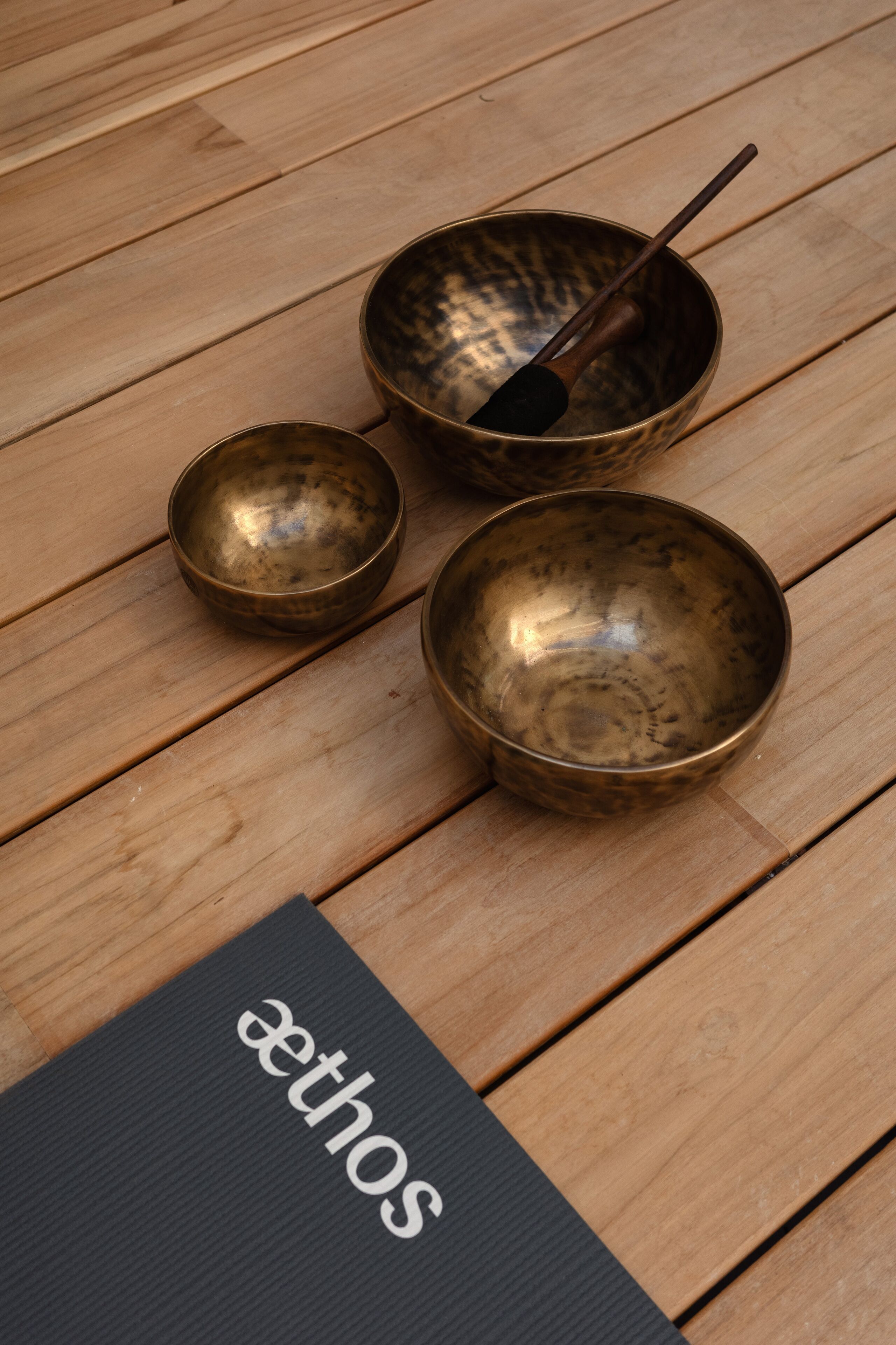 Trois bols en laiton sont posés sur un plancher en bois, sur lequel se trouve un tapis noir portant l’inscription « ethos ».