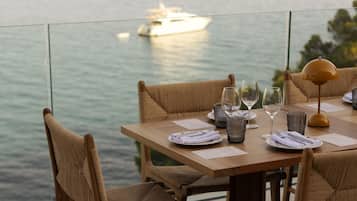 A dining table with wicker chairs, set for four, overlooking a calm sea with a boat in the distance.
