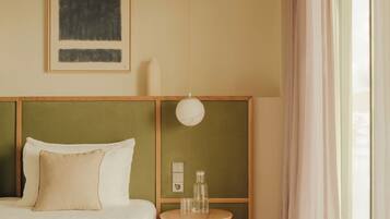 Une chambre d’hôtel moderne avec un lit, une petite table d’appoint ronde en bois, un luminaire suspendu et une grande fenêtre avec des rideaux.
