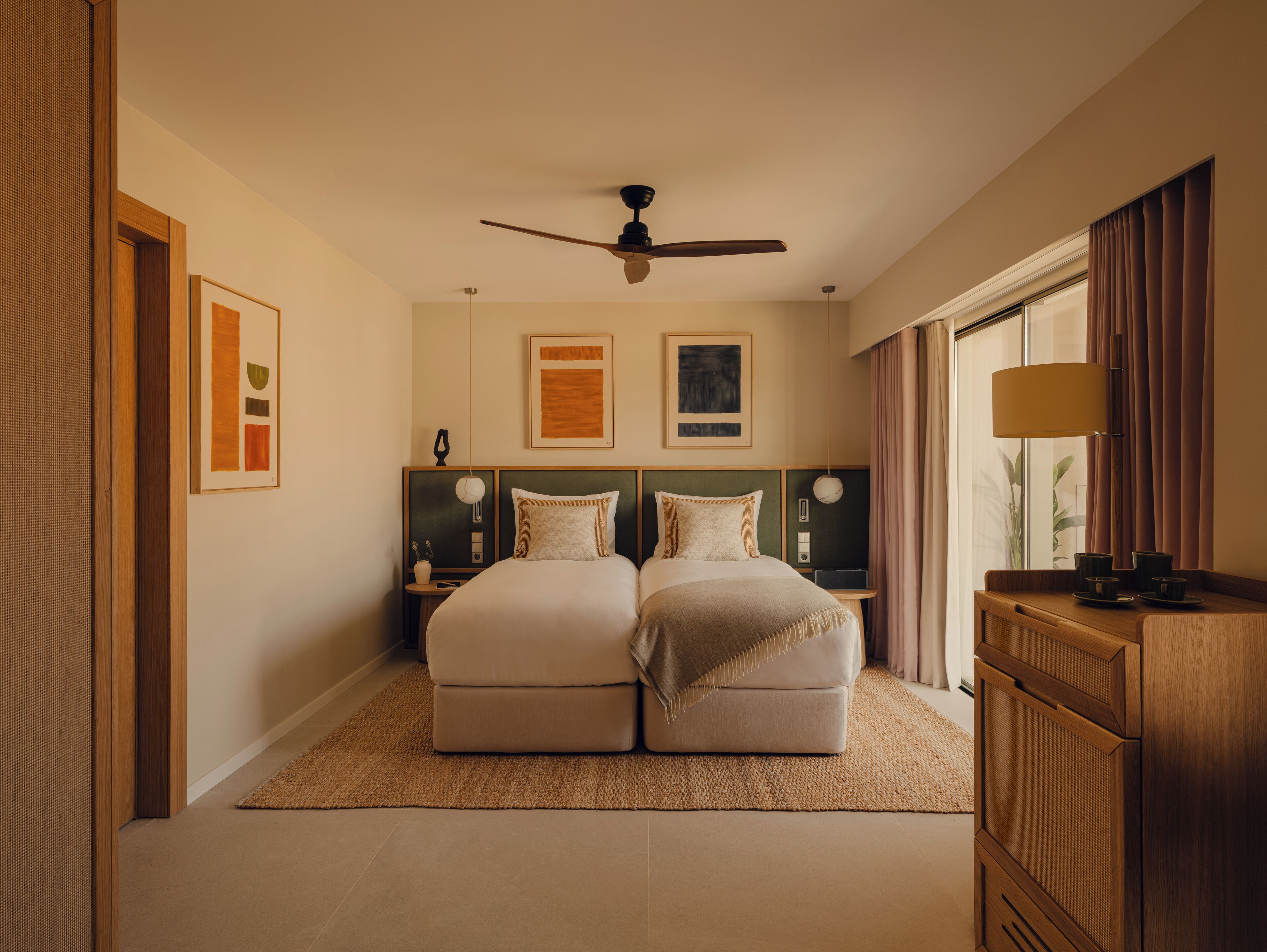 Une chambre d’hôtel avec deux lits, un ventilateur de plafond, des œuvres d’art accrochées aux murs, une table de chevet avec une lampe et une vue sur l’extérieur par une fenêtre.