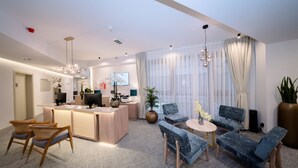 Een moderne hotellobby met een receptiebalie, een zithoek en grote ramen voorzien van dunne gordijnen.