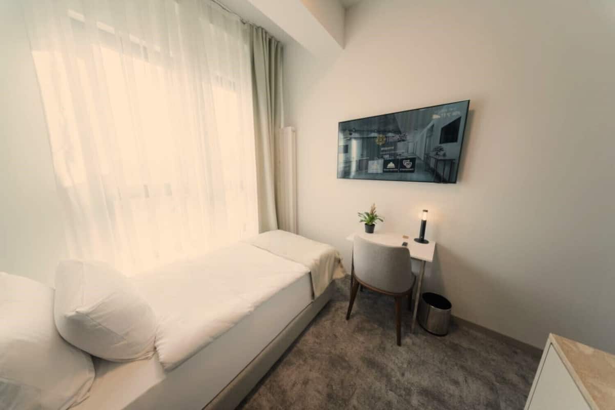 Ein kleiner, gut beleuchteter Raum mit einem Einzelbett, einem Schreibtisch mit Stuhl, einer Topfpflanze und einem gerahmten Bild an der Wand.