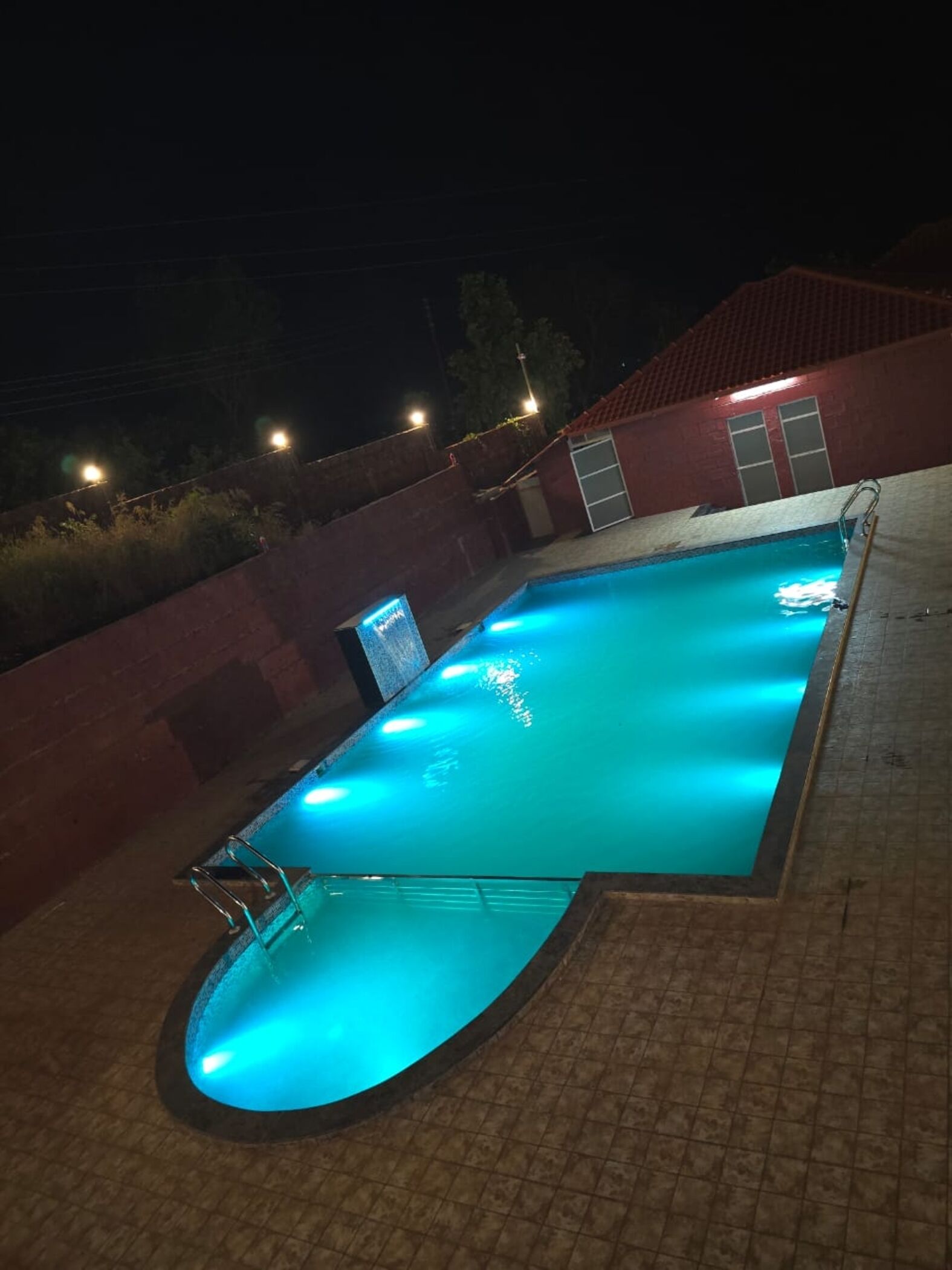 Una piscina con borde de baldosas, una escalera y un edificio con techo rojo al fondo.