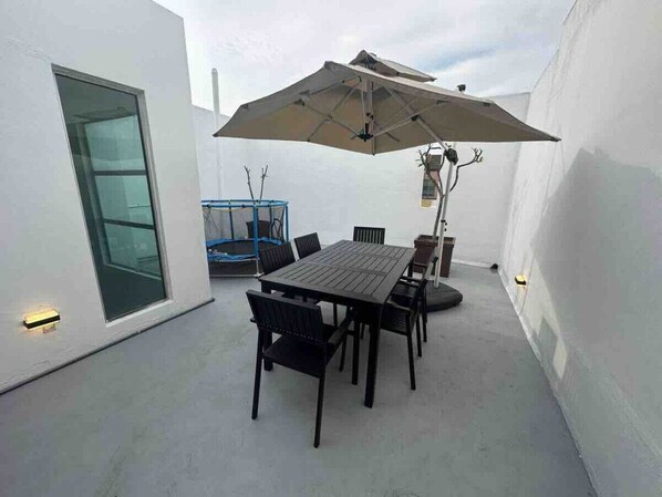 An outdoor patio with a table and chairs, a large umbrella, and a blue trampoline.