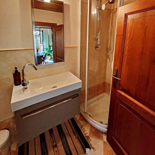 A bathroom with a white sink, a mirror above it, a shower with a glass door, and a wooden door. - The Old Washhouse (Radicofani)