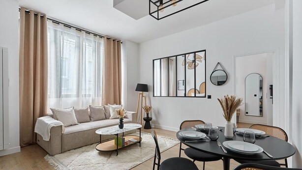 Una sala de estar moderna con sofá, mesa de centro, mesa de comedor y sillas. Hay un ventanal con cortinas, una lámpara y una obra de arte en la pared.