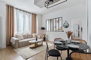 Uma sala de estar moderna com sofá, mesa de centro, mesa de jantar e cadeiras. Há uma janela grande com cortinas, uma luminária e uma obra de arte na parede.
