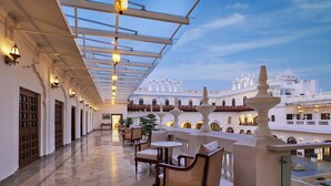 Un balcón espacioso con techo de cristal, amueblado con sillas y mesas de mimbre, con vistas a un patio con edificios blancos y una fuente.