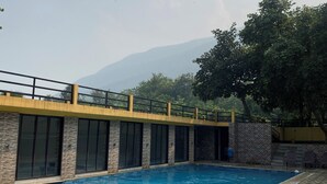 A swimming pool with a tiled deck, a covered area with windows, and a view of mountains in the background. - Panchet Residency (Raghunathpur)