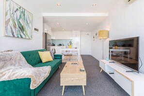 A modern living room with a green sofa, a wooden coffee table, a flat-screen TV, and a kitchen in the background.