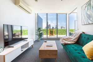 A modern living room with a large flat-screen TV, a wooden coffee table, a green sofa, and a large window offering a city view.