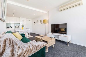 A modern living room with a green sofa, a wooden coffee table, a TV, and a kitchen in the background.