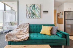 A teal sofa with a furry throw blanket, a green wall with a framed abstract painting, a white door, and a stainless steel refrigerator.