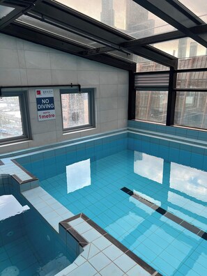An indoor swimming pool with tiled walls and windows, a 'NO DIVING 1 metre' sign, and a view of buildings outside.