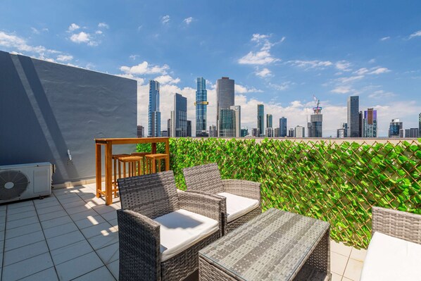 A rooftop terrace with wicker furniture, a fan, and a view of a city skyline.