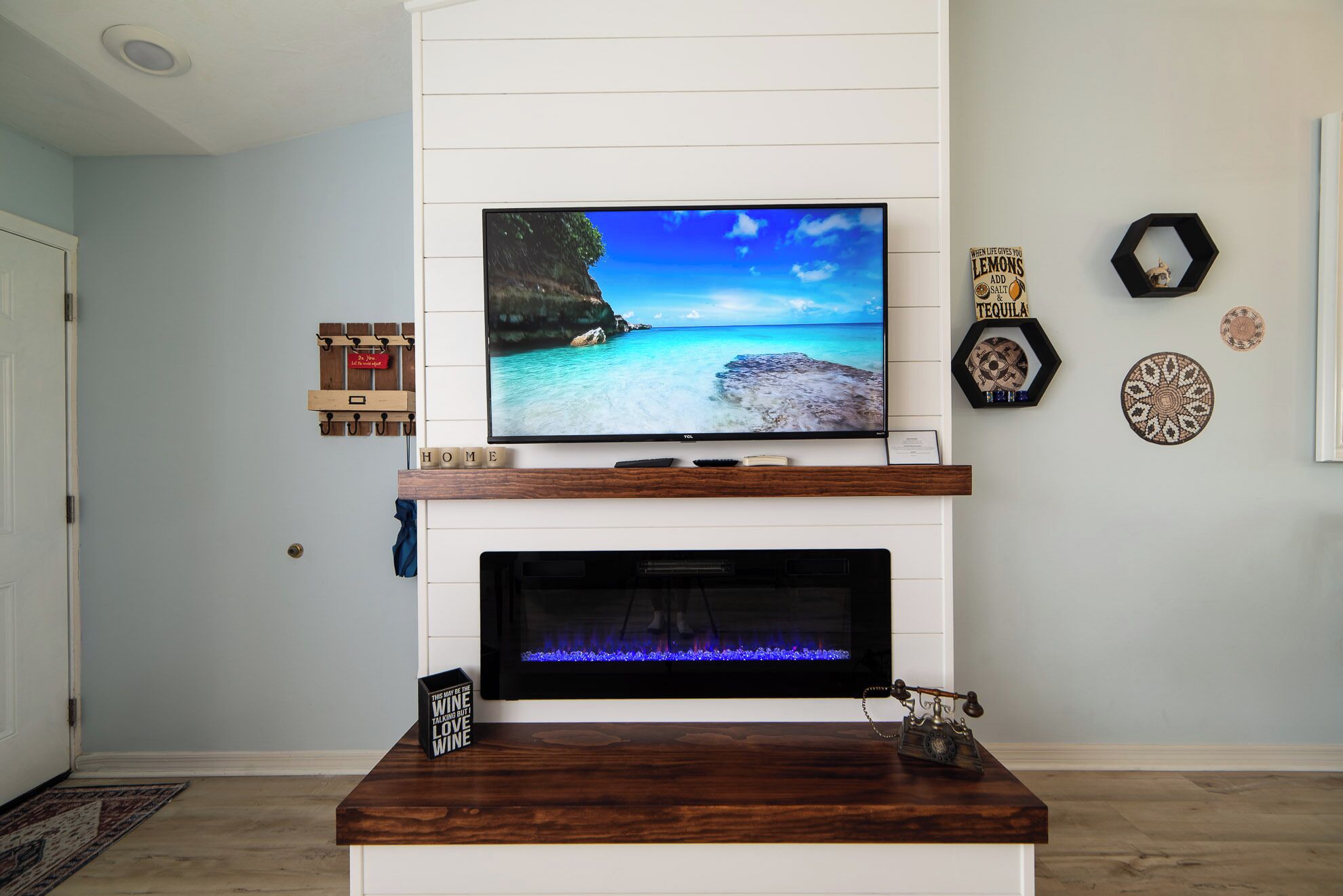 Een woonkamer met een flatscreen televisie boven een open haard, een wandtelefoon en decoratieve muurkunst.
