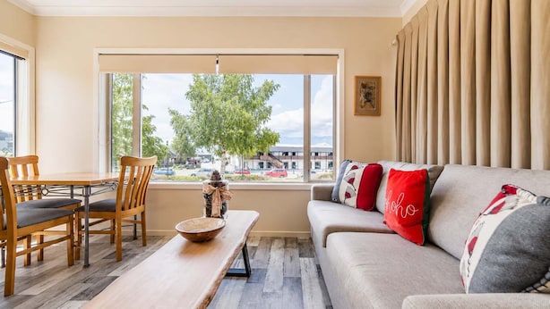 A living room with a sofa, chairs, a dining table, and a large window with a view of a tree and buildings. - Starlight Suite | Tekapo 1BR + Lake Walk (Lake Tekapo)