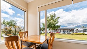 A dining area with a table and chairs, large windows with a view of a parking lot and buildings, and a balcony with a railing. - Starlight Suite | Tekapo 1BR + Lake Walk (Lake Tekapo)
