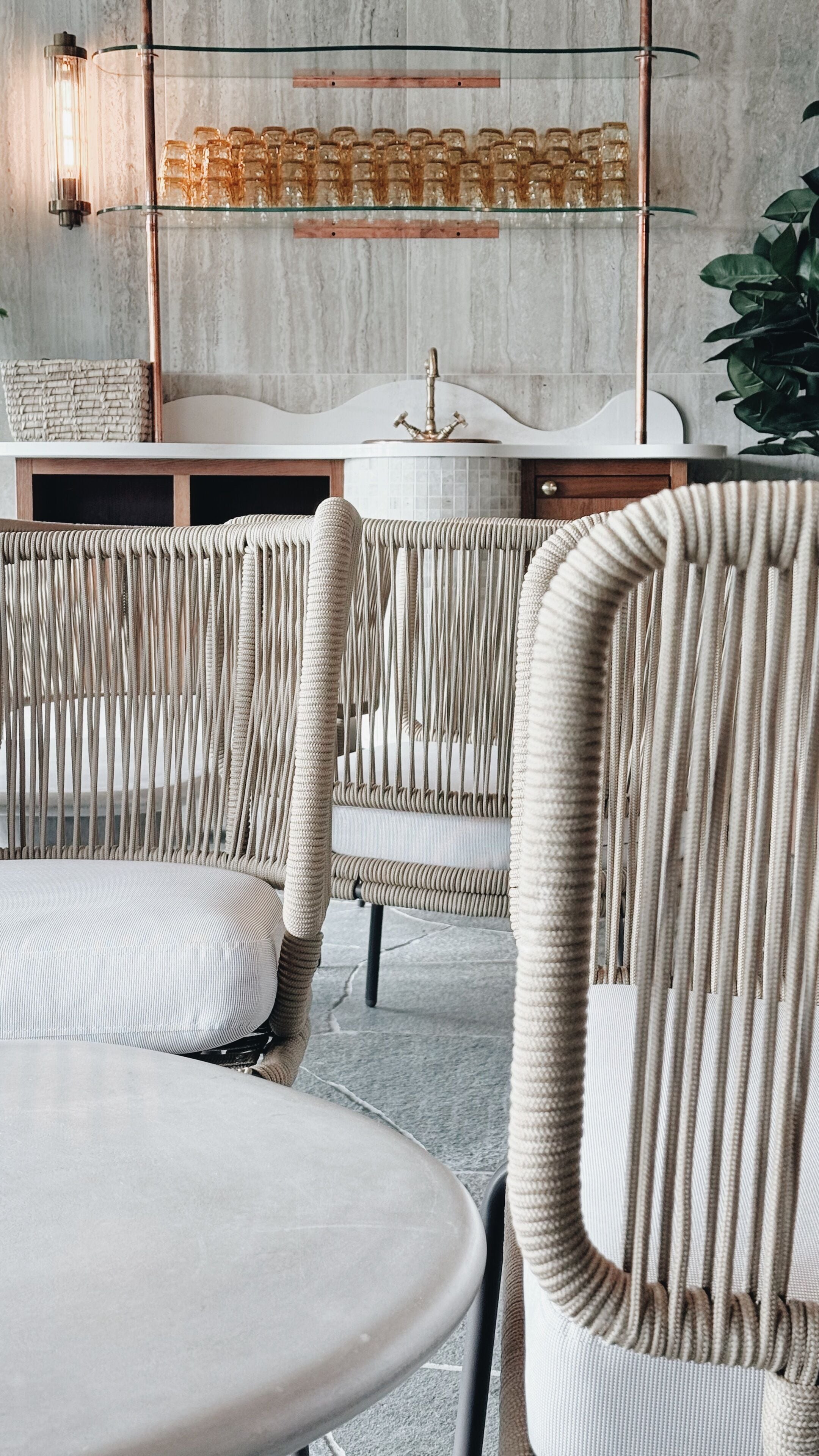 A modern interior with a round dining table, two high-back chairs with vertical slats, a glass shelf with decorative items, and a white marble wall.