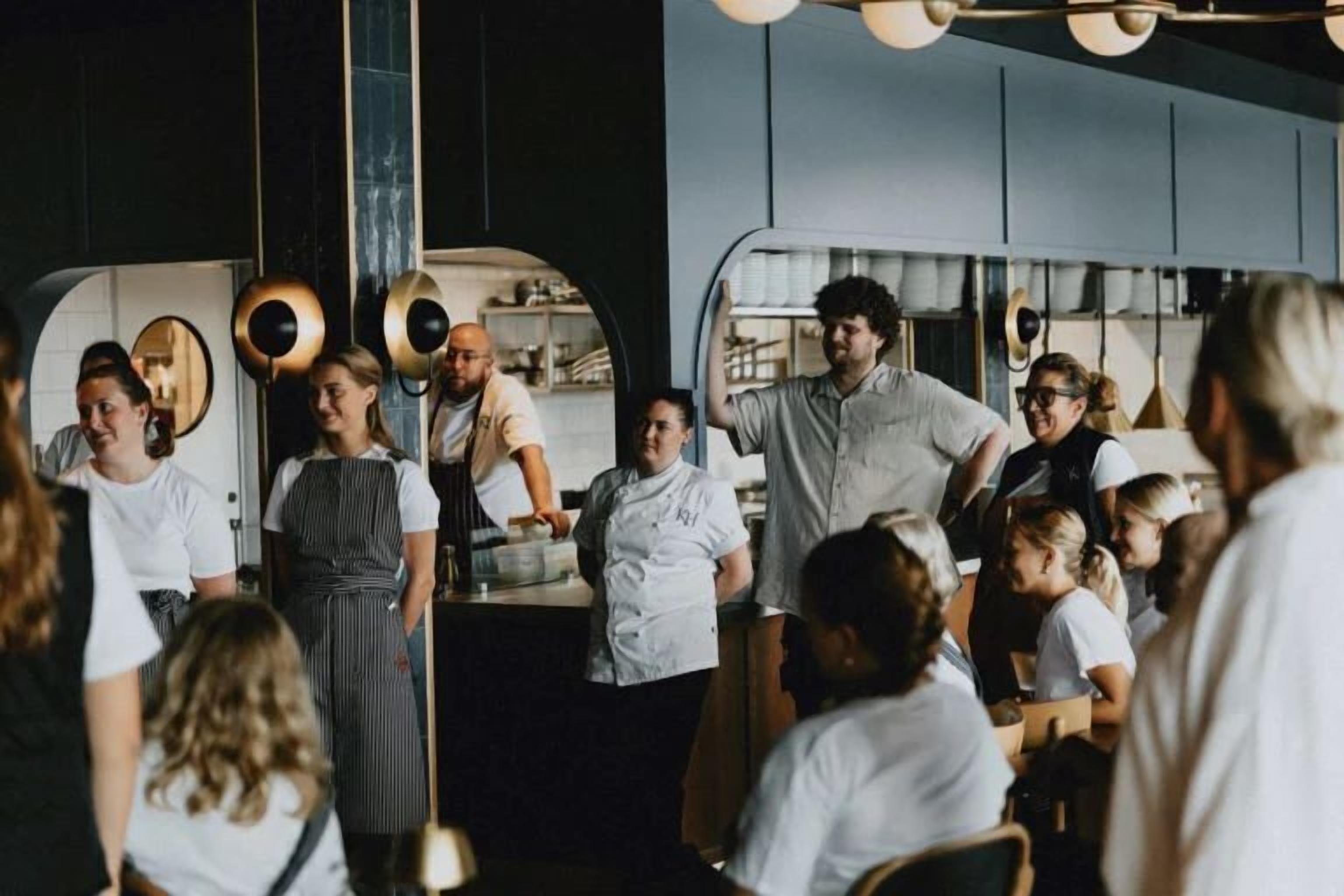 A group of people in a restaurant setting, some standing and some seated, with a counter and kitchen area visible in the background.