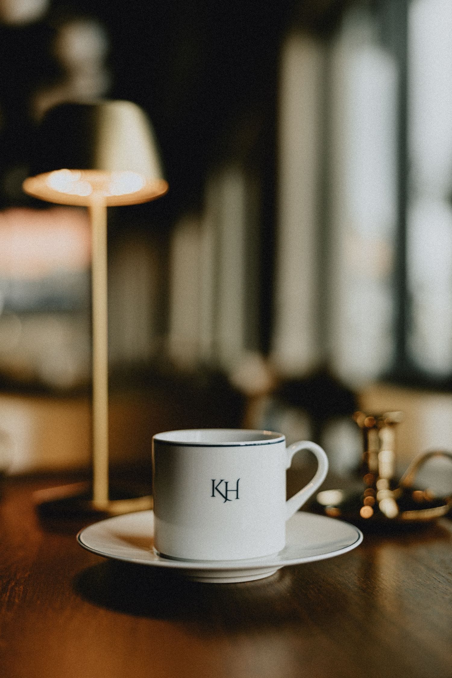 Una taza blanca con la inscripción "KH" sobre un platito, una lámpara de mesa y un fondo desenfocado.
