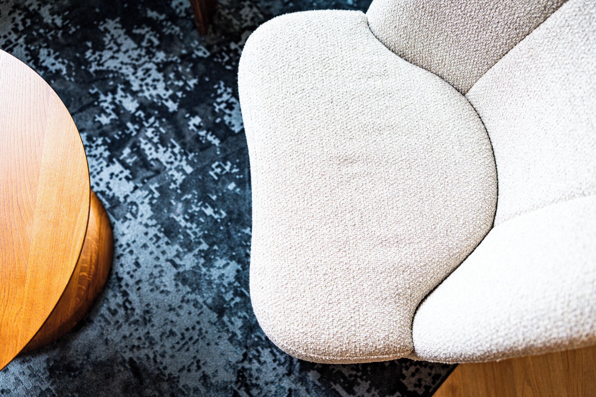 Primer plano de un sillón tapizado en beige con tela texturizada, ubicado junto a una mesa redonda de madera sobre una alfombra estampada.