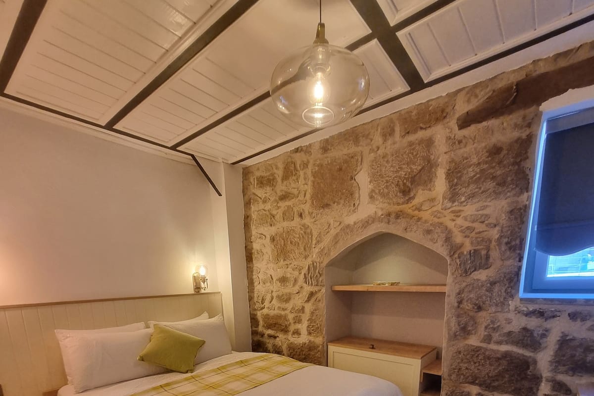 Un dormitorio con pared de piedra, cama con manta a cuadros blanco y amarillo, mesita de noche de madera, silla de madera y televisor montado en la pared de piedra.