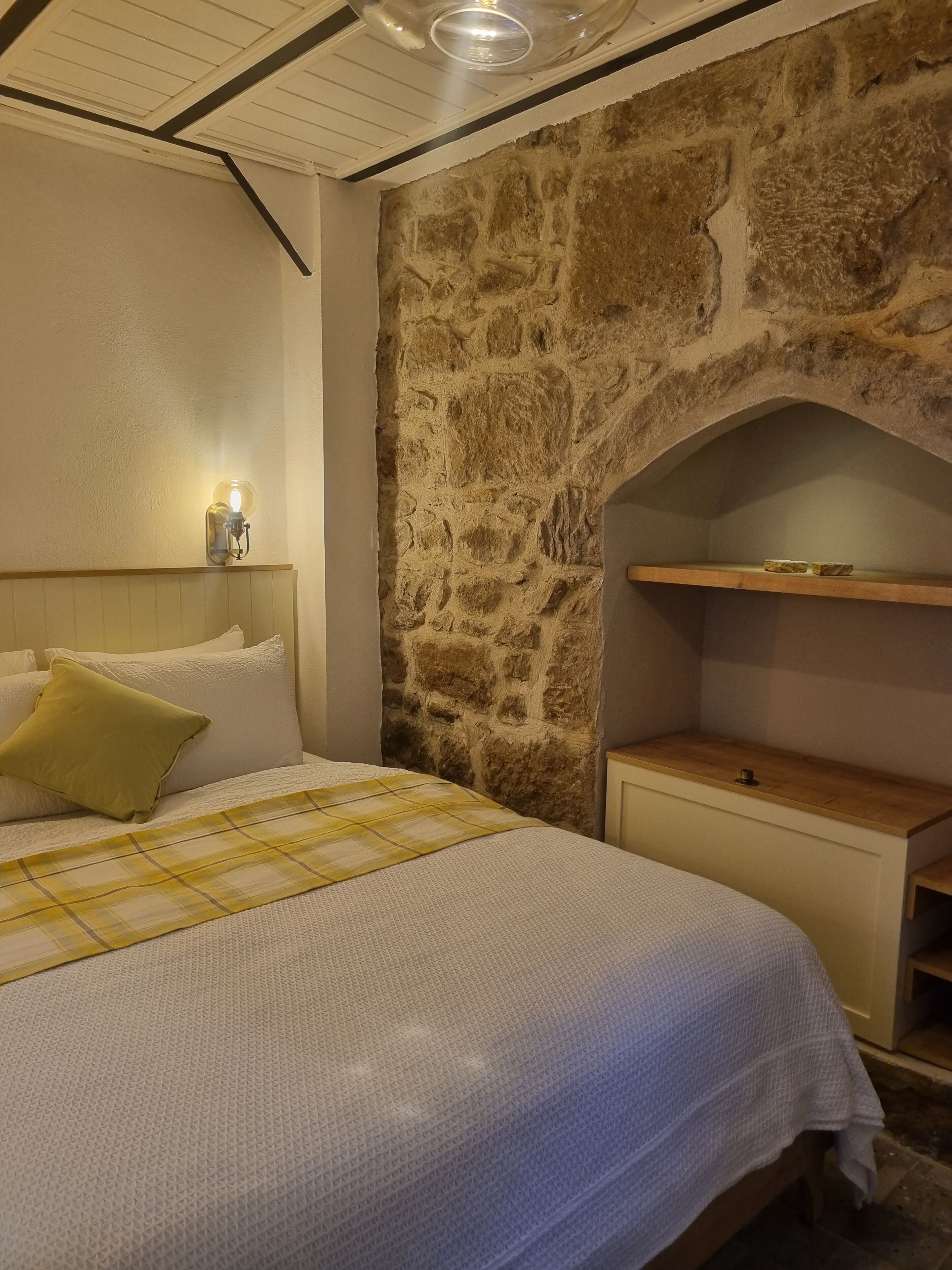 Ein Schlafzimmer mit Steinwand, einem Bett mit gelb-weißer Schachbrettdecke, einem Nachttisch und einer Wandleuchte.