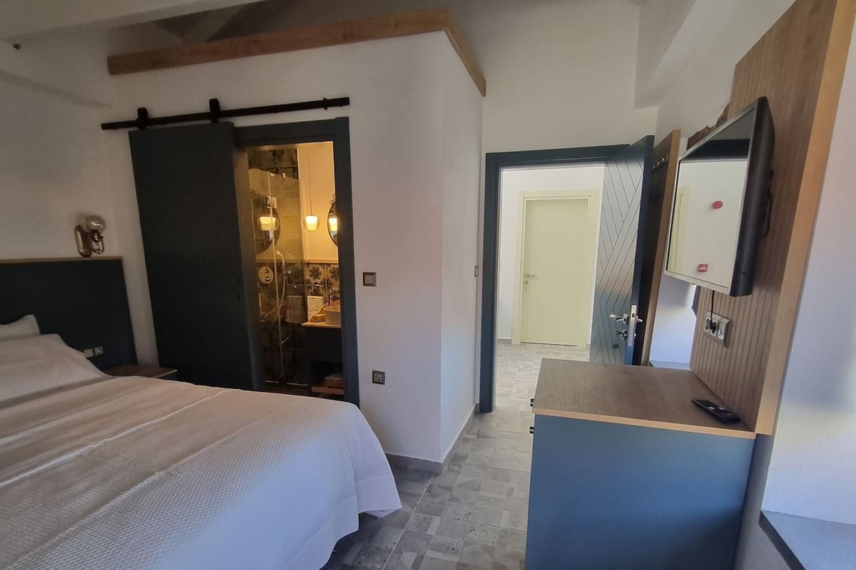 Habitación de hotel con cama, escritorio, silla, espejo y baño visible a través de una puerta abierta.