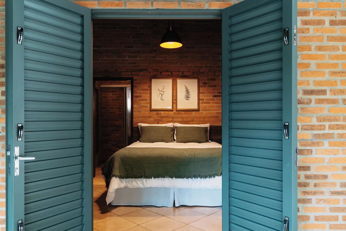 Un dormitorio con pared de ladrillo, una cama con cubrecama verde y blanco, dos grabados botánicos enmarcados y una lámpara colgante.