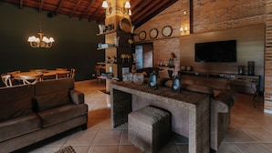 A cozy living area with a wooden ceiling, a brick wall, a TV mounted on the wall, and a dining table with chairs. - Quinta de Brumado (Óleo)
