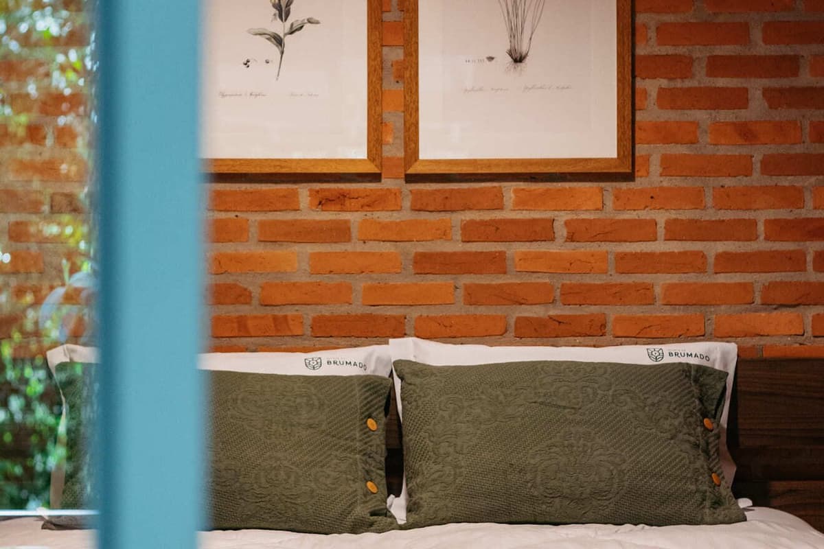 Un dormitorio con pared de ladrillo, dos ilustraciones botánicas enmarcadas, cama con ropa de cama verde y ventana con vistas a vegetación.