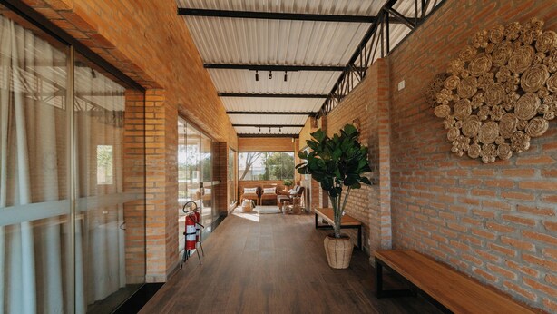 A corridor with wooden flooring, brick walls, and a ceiling with metal beams. There is a large potted plant and a decorative wall piece. - Quinta de Brumado (Óleo)