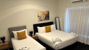 Hotellihuoneessa on kaksi sänkyä, joissa kummassakin on valkoiset lakanat ja yksi ruskea tyyny. Seinällä on taulu, sängyn vieressä on yöpöytä, jossa on lamppu, ja ikkunassa on valkoiset verhot.