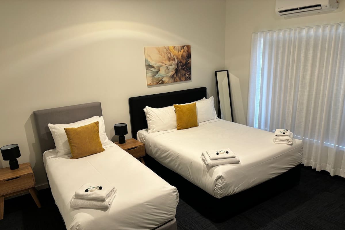 Une chambre d’hôtel avec deux lits, chacun étant équipé de draps blancs et d’un seul oreiller marron. Un tableau est accroché au mur, il y a une table de chevet avec une lampe, et une fenêtre avec des rideaux blancs.